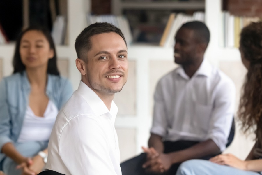 Portrait of smiling Caucasian man look at camera sit in circle at group psychological counselling, relieved male posing for picture undergo treatment at therapy session. Sensitivity training concept